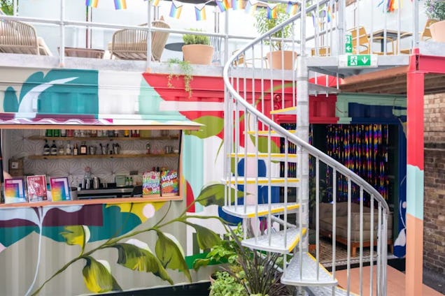 Vibrant outdoor space featuring shipping containers with murals, a spiral staircase, a rooftop terrace, and a bar area with colourful bunting.