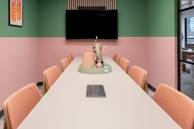 Meeting room with a long white table, peach chairs, and two-tone green and pink walls beneath a wall-mounted screen.