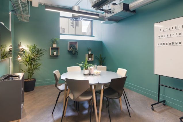 Meeting room with a round grey table, teal walls, industrial ceiling pipes, and a whiteboard standing on a mobile base.
