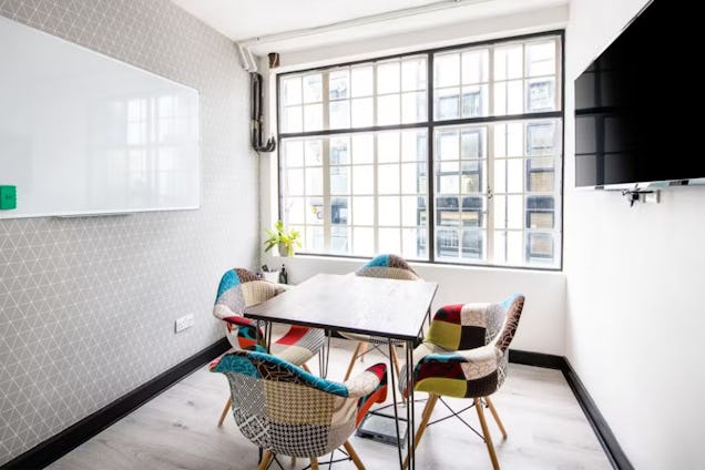 Compact meeting room with a dark square table, colourful patchwork chairs, a large industrial window, and patterned grey wallpaper.