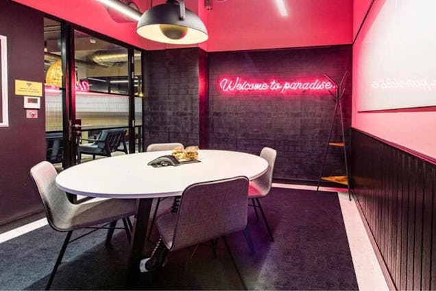Meeting room with a round white table, grey chairs, and a pink neon sign reading "welcome to paradise" on a black textured wall.