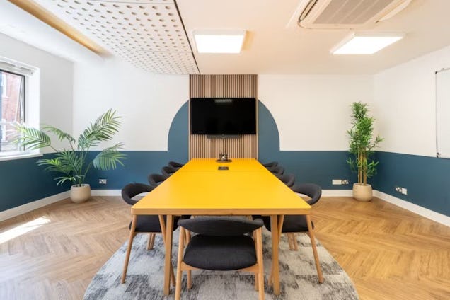 Meeting room with a vibrant yellow table, teal-and-white walls, herringbone flooring, and a slat-wood feature wall behind a screen.