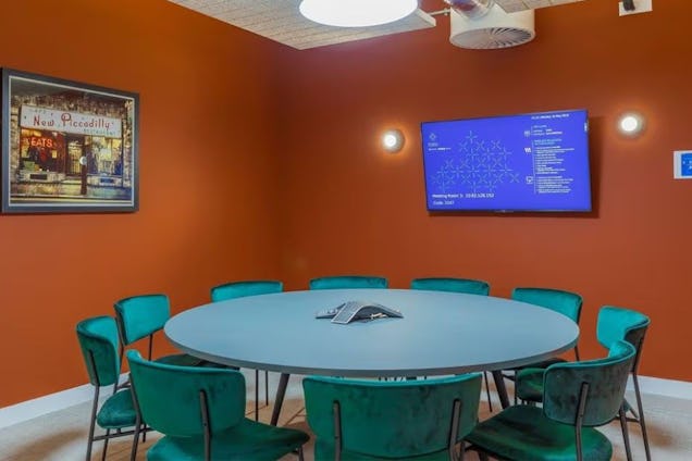 Meeting room with a round grey table, teal velvet chairs, burnt orange walls, and a wall-mounted screen displaying data.