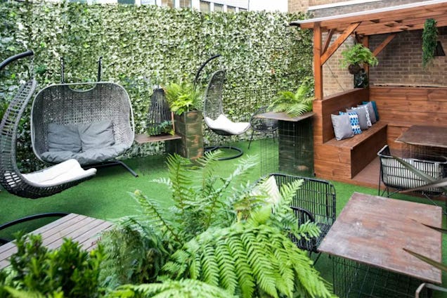 Outdoor terrace with artificial grass, featuring woven egg chairs, wooden bench seating under a pergola, and green privacy screening.
