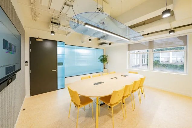 Bright meeting room with an oval wood table, yellow chairs, light blue glass partitions, and a unique curved transparent ceiling panel.