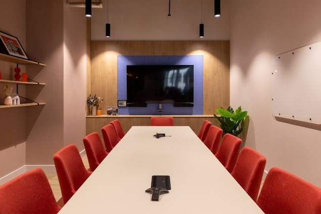 Meeting room with a long white table, orange chairs, and a wood-panelled feature wall with a blue-framed TV.