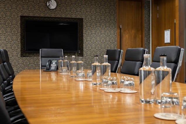 Executive boardroom with a long wooden table, black leather chairs, water jugs, and a patterned feature wall with a large screen.