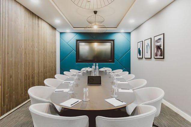 Boardroom with an oval table, white tub chairs, a wood-slat wall, and a teal geometric feature wall with a mounted screen.