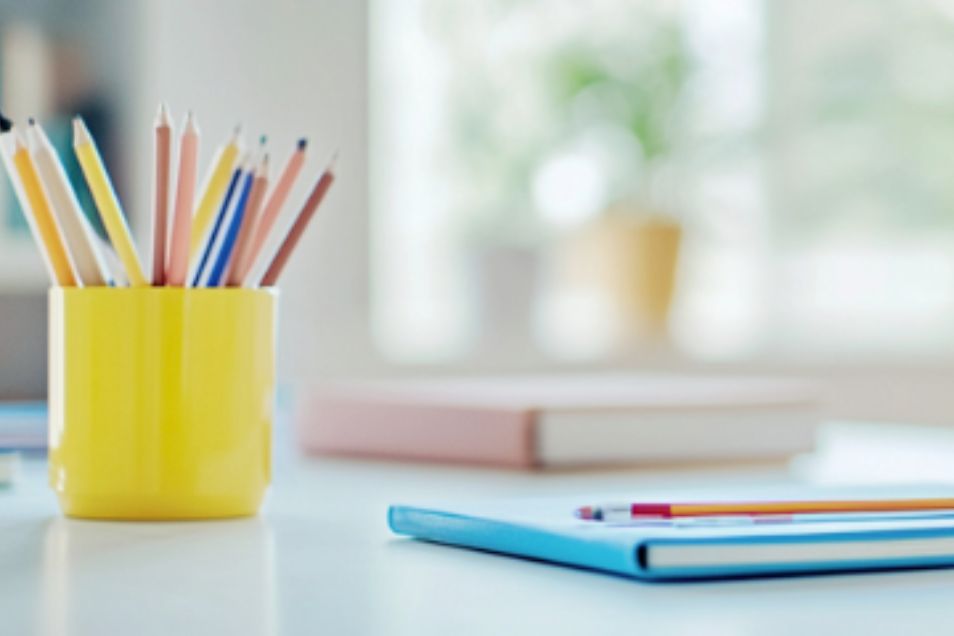 Yellow cup holding coloured pencils beside notebooks on a bright desk with a softly blurred window background.