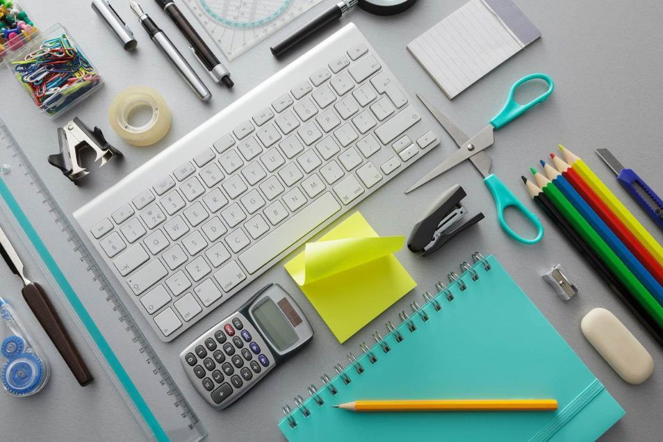 Top-down view of a desk with keyboard, calculator, sticky notes, pens, scissors and neatly arranged stationery on a grey surface.
