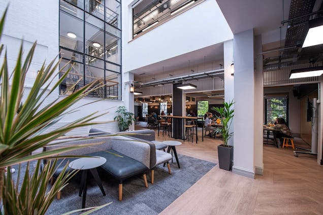 Office lounge with sofas, tables and greenery beneath a glass mezzanine, leading into an open workspace.