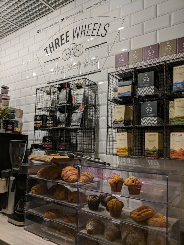 Bakery counter with pastries in a glass display, coffee equipment and shelves of products against tiled walls.