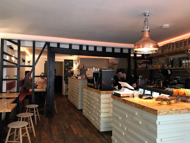 Warmly lit café interior with wooden counters, stools and a display of pastries, with staff serving behind the bar.