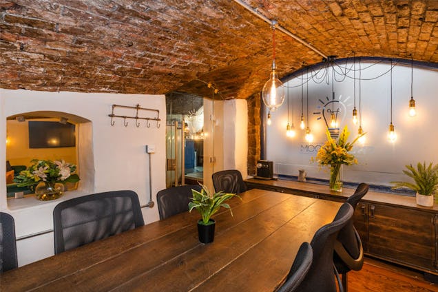 Meeting room with a long wooden table, exposed brick ceiling, glass partition and warm pendant lighting.