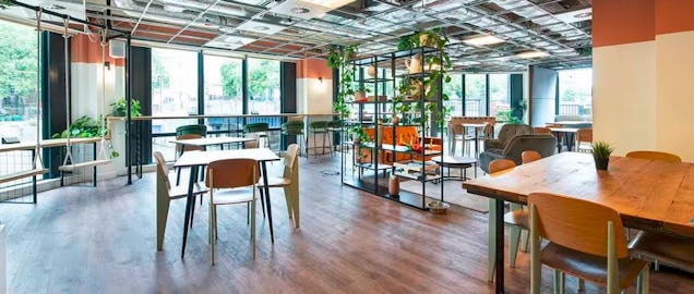 Bright open workspace with wooden tables, large windows and greenery, offering a modern communal working area.
