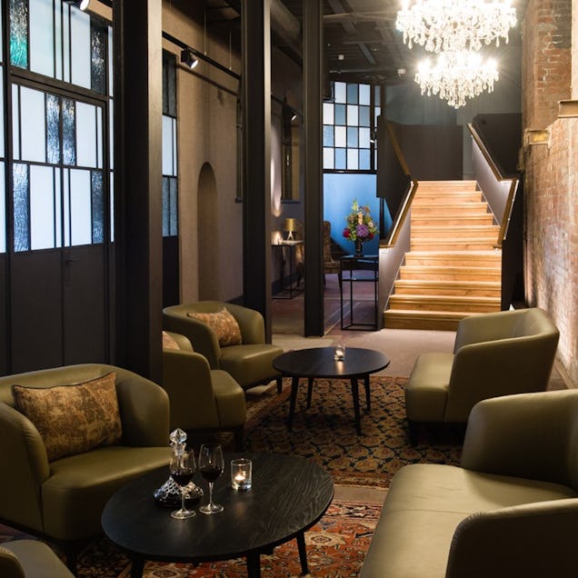 Lounge with armchairs, chandeliers and a patterned rug, leading to a staircase in a softly lit interior.