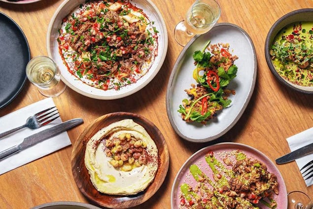 Assorted dishes, including salads, hummus and grain bowls, arranged on a table, with drinks and cutlery nearby.