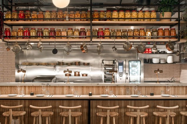 Bar area with wooden stools, a long counter and shelves lined with jars, bottles and glassware.
