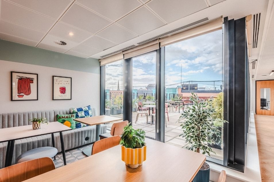 Sunlit office break room with wooden tables and a cushioned bench, featuring large glass doors that open onto an outdoor terrace.