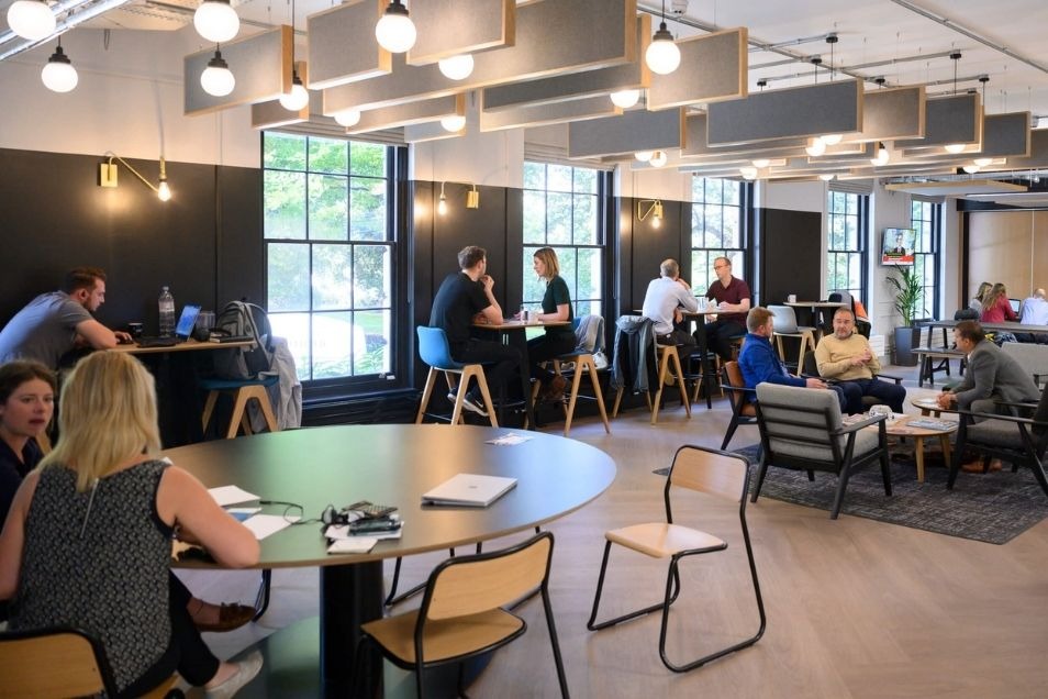 Coworking space with grey acoustic ceiling baffles, globe lights, and people working at round tables and window-side desks.