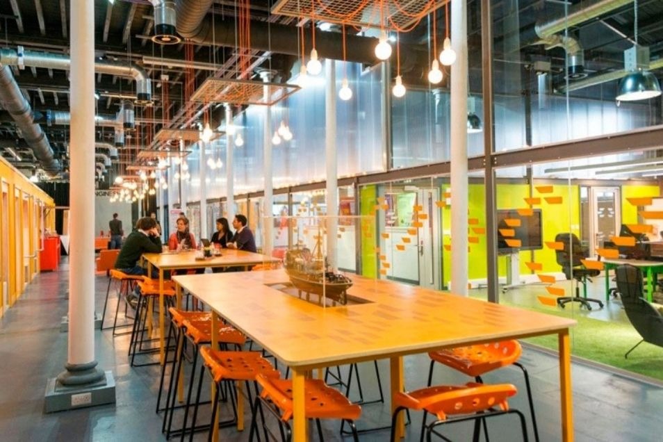 Office with long wooden communal tables, orange stools, and hanging bulbs set against glass-walled meeting rooms.