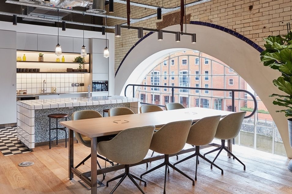 Office kitchen and dining area featuring a large arched window, a tiled island, and a long wooden table with cream chairs.