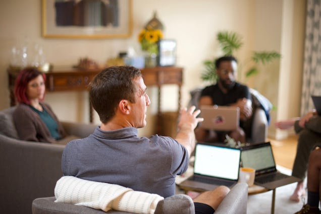 A man gestures while leading a group discussion in a lounge with people sitting in grey armchairs with laptops.
