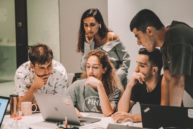 A group of five young people lean in with focused expressions to look at a laptop screen together on a white desk.