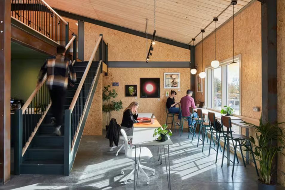 Coworking space with plywood walls, high ceilings, and people working at desks or window-side seating near a staircase.