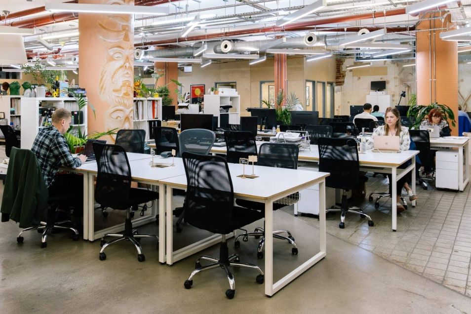 Open-plan office with white communal desks, black mesh chairs, and terracotta-coloured pillars under exposed ceiling pipes.
