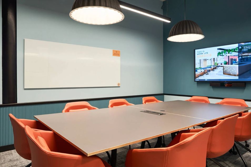 Meeting room with a large rectangular table, orange armchairs, a whiteboard, and a wall-mounted screen under pendant lighting.