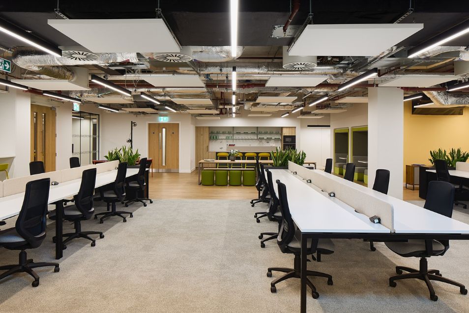 Open-plan office with white workstations, black ergonomic chairs, and a complex industrial ceiling featuring acoustic panels and exposed ductwork.