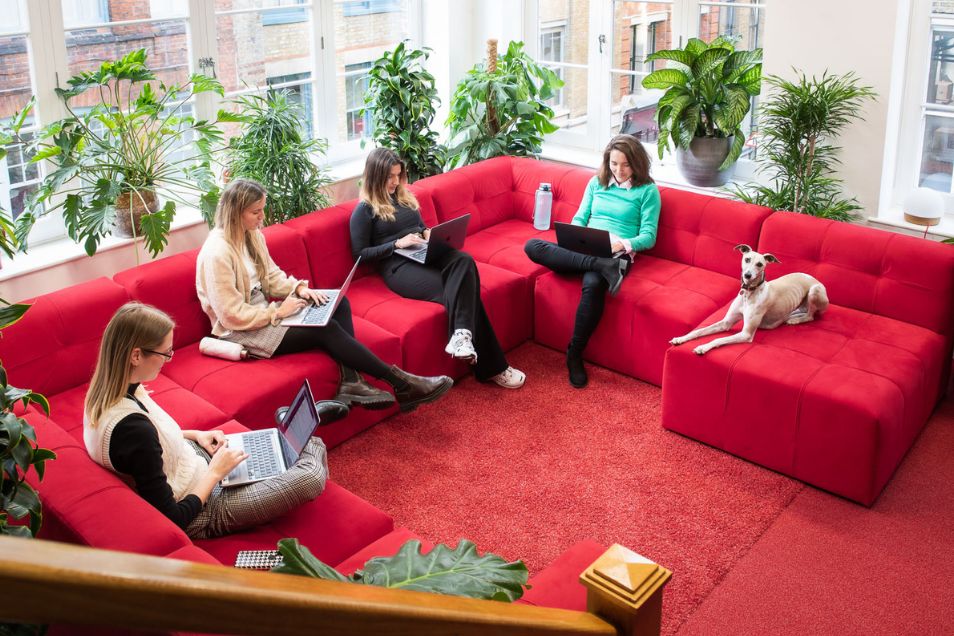 Four people and a dog relax on a large, U-shaped red modular sofa in a bright, plant-filled office lounge.