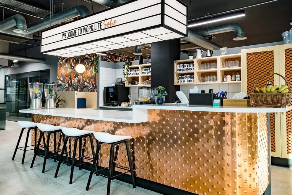 Office bar with a textured copper-fronted counter, white bar stools, and a large grid-style light fixture overhead.