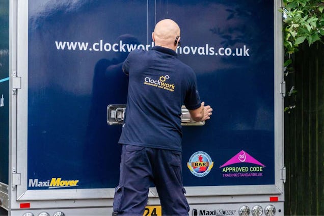 A mover in a navy uniform closing the back of a blue Clockwork Removals van.