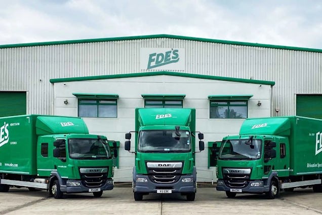 Three green Ede’s moving trucks parked in front of a large white warehouse.