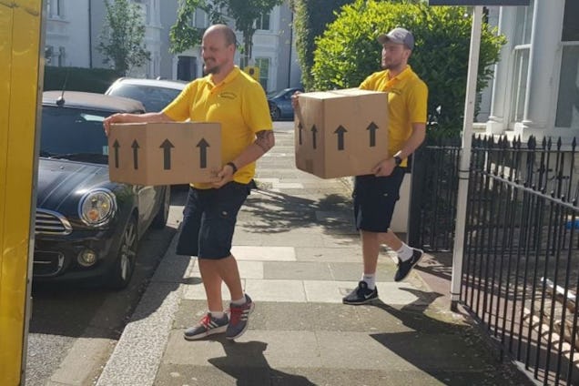 Two movers in yellow shirts carrying large cardboard boxes along a residential sidewalk.