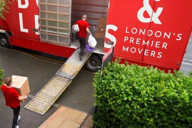 Movers in red shirts use a ramp to load boxes into a large red truck labeled "London's Premier Movers."