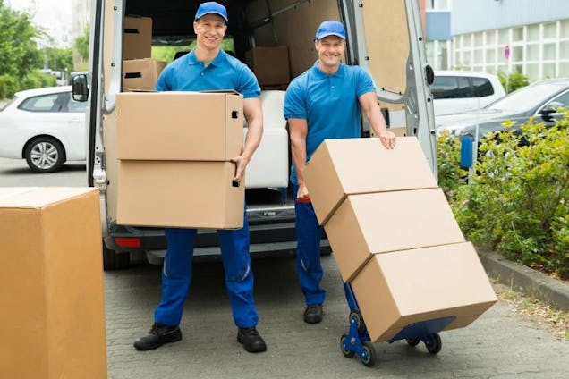 Two smiling movers in blue uniforms loading large cardboard boxes from a van.