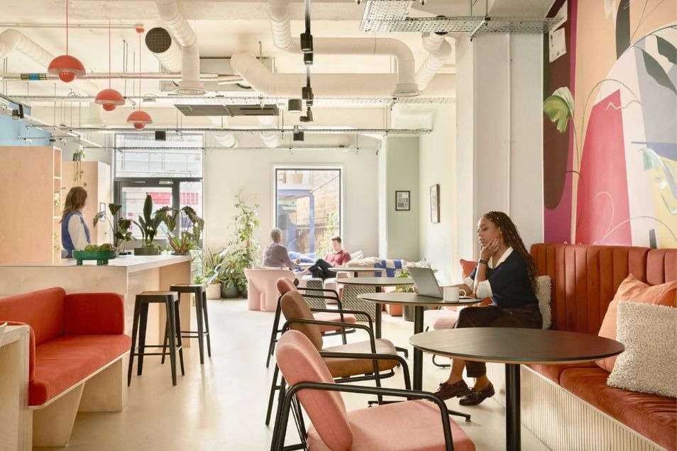 People work and socialise in an open-plan office cafe featuring pink armchairs, a long seating chair, and industrial lighting.