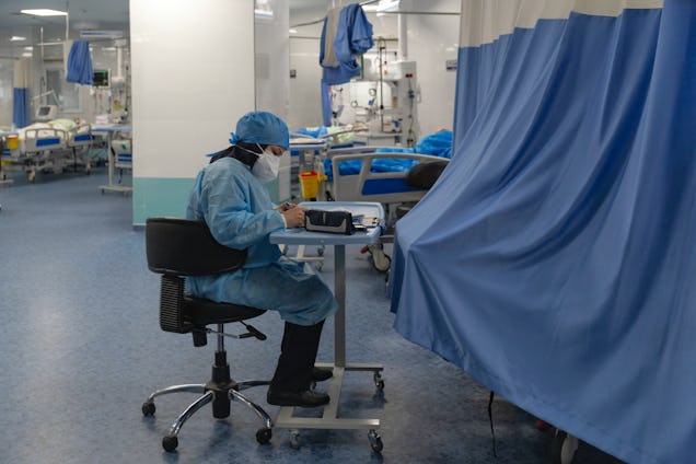 A medical professional in full blue protective gear sits and works in a large hospital ward.