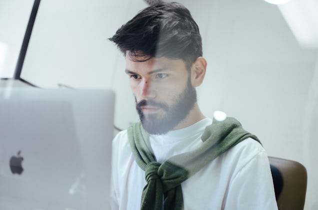 A man with a sweater draped over his shoulders works intently on a laptop in a bright indoor setting.