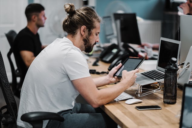 A man with a bun sits at a wooden desk in a busy office and checks a tablet while working on a laptop.