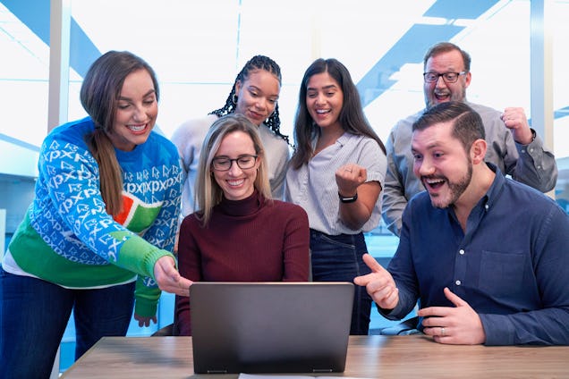 A group of diverse colleagues gathered around a laptop, smiling and celebrating a success in a bright office.