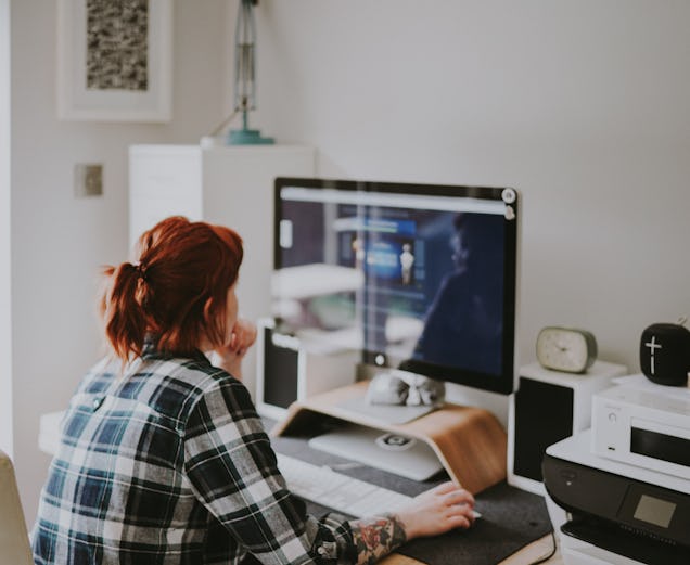 A woman in a flannel shirt sits at a desk and works on a desktop computer in a home office.