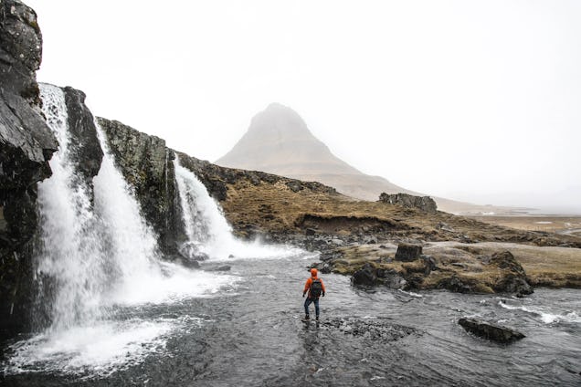 A hiker in an orange jacket stands in a shallow river near a powerful waterfall with a misty mountain in the background.