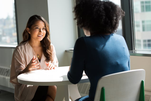 Two women sit at a small white circular table in an office setting and engage in a focused conversation.