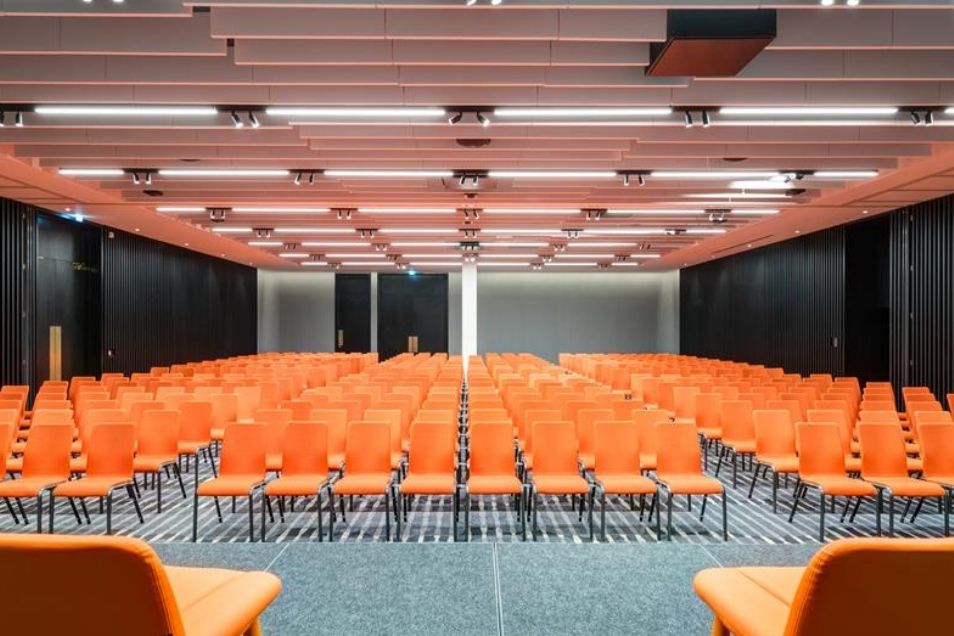 A large, modern auditorium filled with neat rows of orange chairs facing a wide, open floor space.