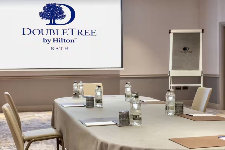 A meeting room at DoubleTree by Hilton Bath, featuring a projector screen with the hotel logo above a set boardroom table.
