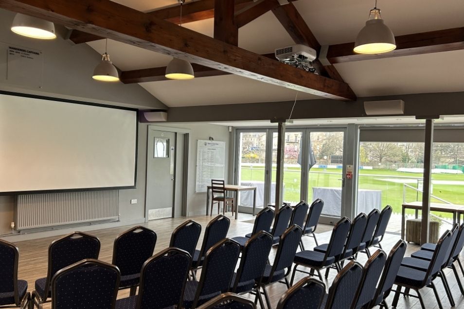 Large presentation room with rows of black chairs facing a projector screen beneath rustic wooden ceiling beams.